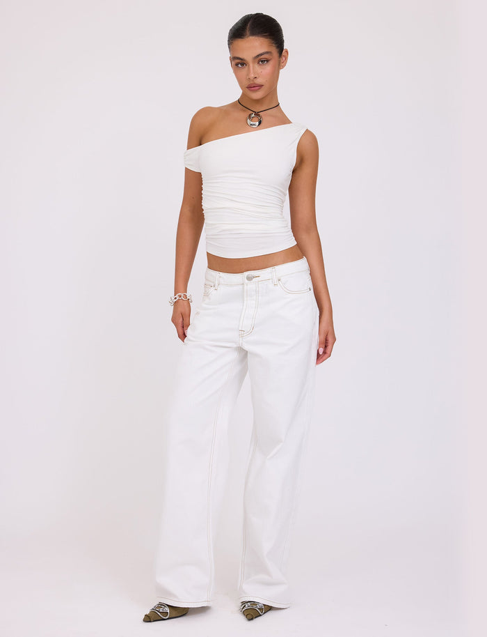 A person stands against a plain white background, wearing the asymmetrical FELIX JEAN - WHITE top and wide-leg jeans from TIGERMIST. They accessorize with a necklace, bracelet, and pointed-toe shoes. Their hair is pulled back as they pose with one hand on their hip.