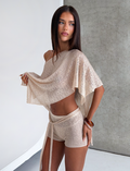 A woman with long brown hair poses by a textured gray wall, wearing the CL - FERN CROCHET SHORT from Tiger Mist USA with a matching sheer, off-shoulder top, holding up her top and gazing to the side neutrally.