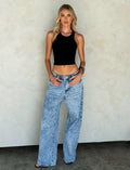 A woman with long blonde hair is standing in front of a light beige wall. She is wearing a black sleeveless top and high-waisted, loose-fit FERN JEAN - BLUE : DENIM BLUE by TIGERMIST. She has one hand in her pocket and is looking directly at the camera with a neutral expression.