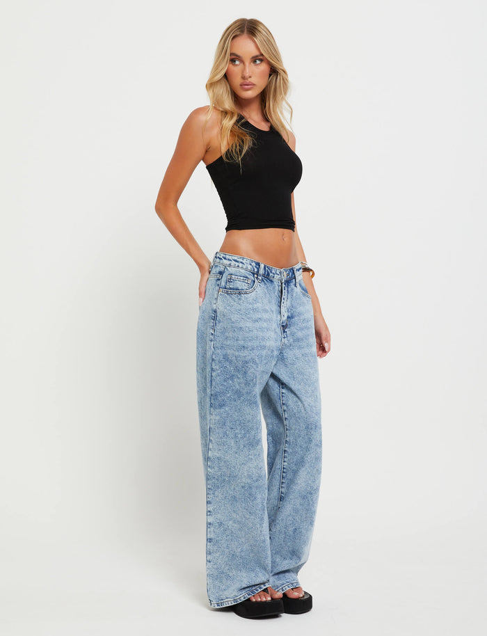 A woman with long blonde hair is wearing a black sleeveless crop top and the TIGERMIST FERN JEAN - BLUE denim jeans. She is posing slightly to the side against a plain white background, with one arm relaxed by her side and the other touching her hair.