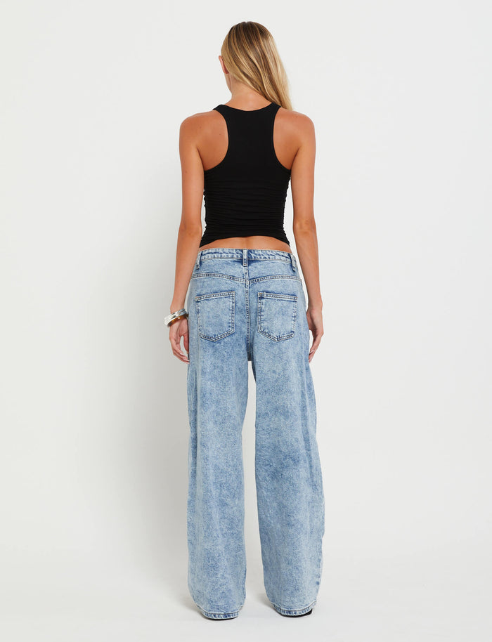A person with long blonde hair stands with their back to the camera against a white background, wearing a black sleeveless top and FERN JEAN - BLUE : DENIM BLUE from TIGERMIST, which are light blue, loose fit denim jeans featuring a high-waisted, wide-leg cut. The person's left wrist is adorned with bracelets.
