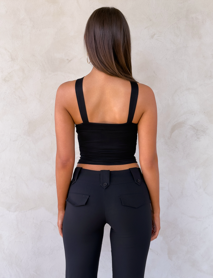 A woman with long brown hair faces a light-textured wall, wearing the TIGERMIST FINCH TOP in black and high-waisted black pants with pocket flaps and visible buttons.