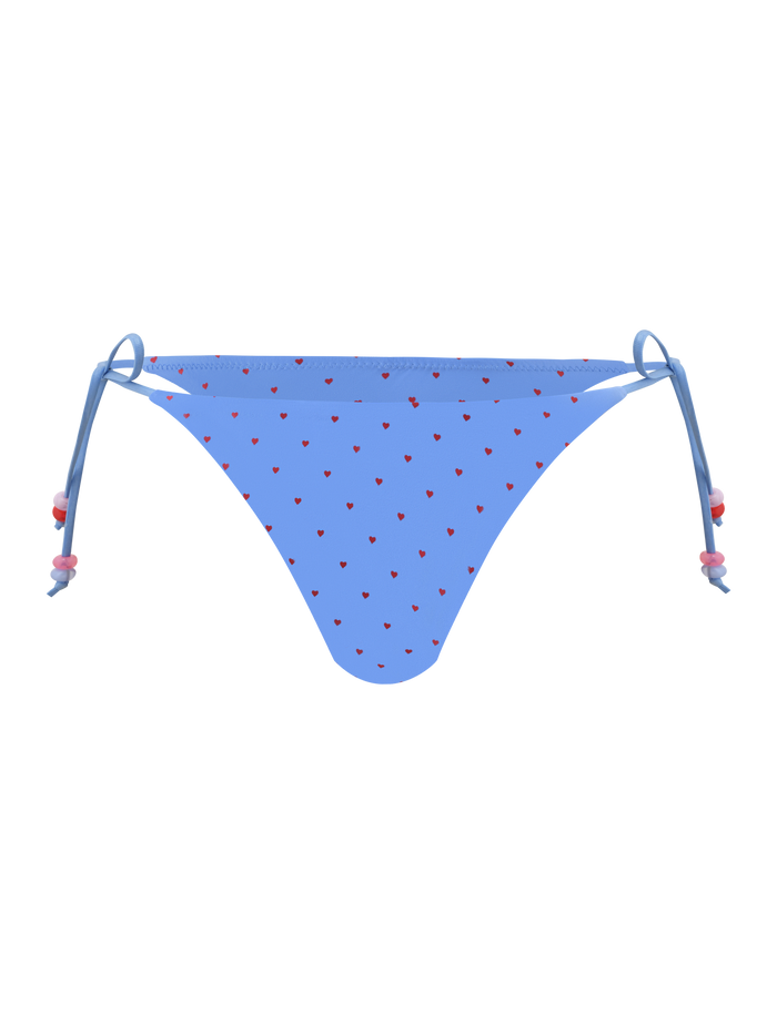 The FINLEA BIKINI BOTTOM - BLUE HEART POLKA DOT by TIGERMIST features a cheeky cut, blue heart print with red hearts, adjustable side ties with pink and red beads, shown on a plain white background.