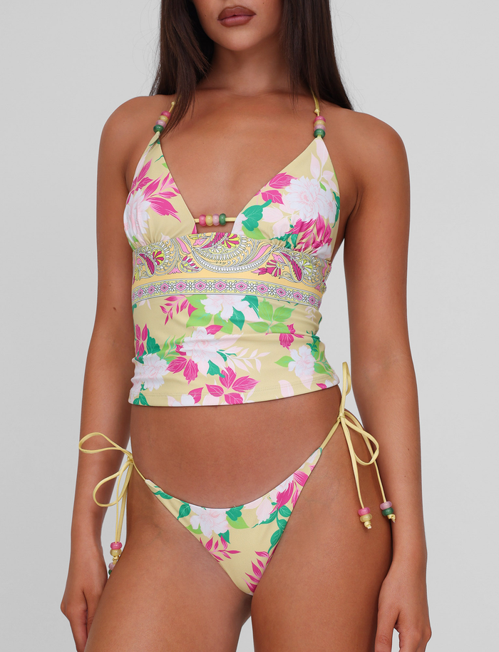 A woman stands against a plain background wearing the TIGERMIST FINLEA BIKINI BOTTOM - PAISLEY BORDER, which features a vibrant paisley print, cheeky cut with side ties and beads, paired with a beaded-strap tankini top.