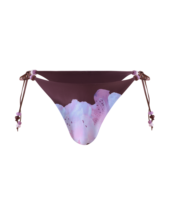 The TIGERMIST FINLEA BIKINI BOTTOM - ZEN FLORAL is a cheeky plum bikini bottom with beaded side-tie straps, featuring soft pink and lavender floral prints on a plain white background.