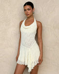 A woman poses against a textured beige wall in the TIGERMIST FIORELLA MINI DRESS—white lace in cream—with a halter neck, draped cowl neckline, and asymmetrical hem. Her dark hair is pulled back and she wears silver hoop earrings.