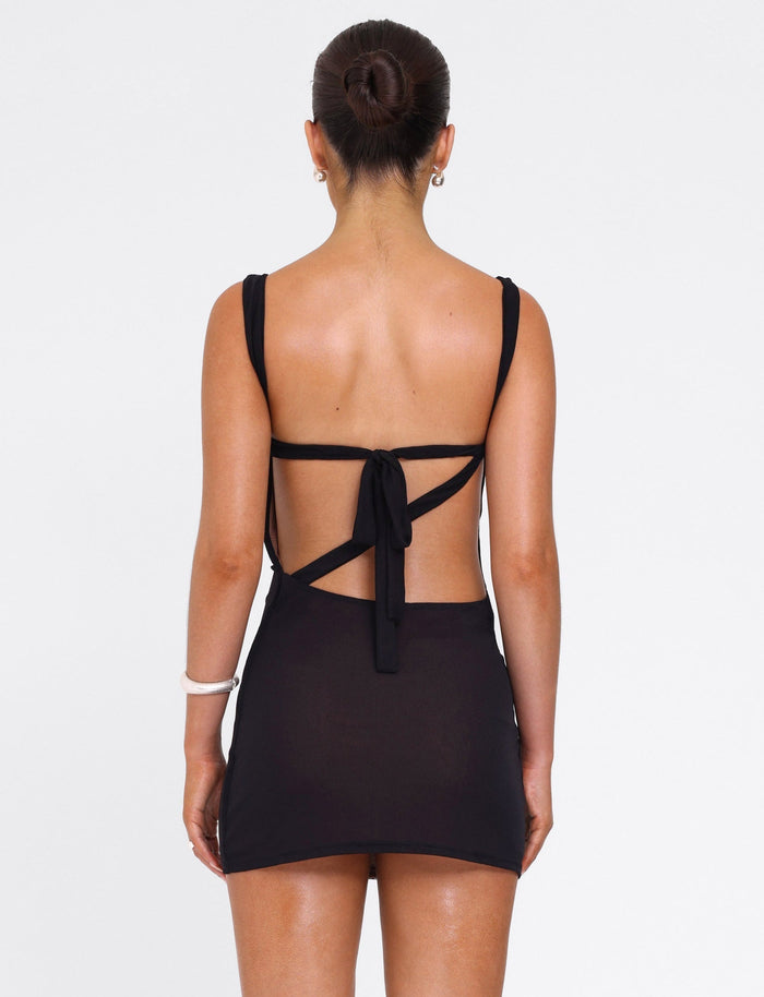 A woman with her hair in a bun wears the TIGERMIST FREYA DRESS - BLACK, a mini dress with an open back and tie detail made from buttery soft fabric, standing against a plain white background.