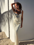 A woman wears the TIGERMIST GINNY MAXI DRESS in off white, standing on sand against a white wall. Sunlight and palm leaf shadows accentuate the draped mesh detail and her elegant silhouette as she gazes to the side.