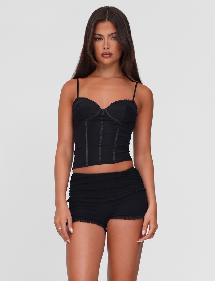 A woman with long brown hair wears the TIGERMIST GINNY SHORT - BLACK and a fitted black camisole, standing against a plain light gray background.