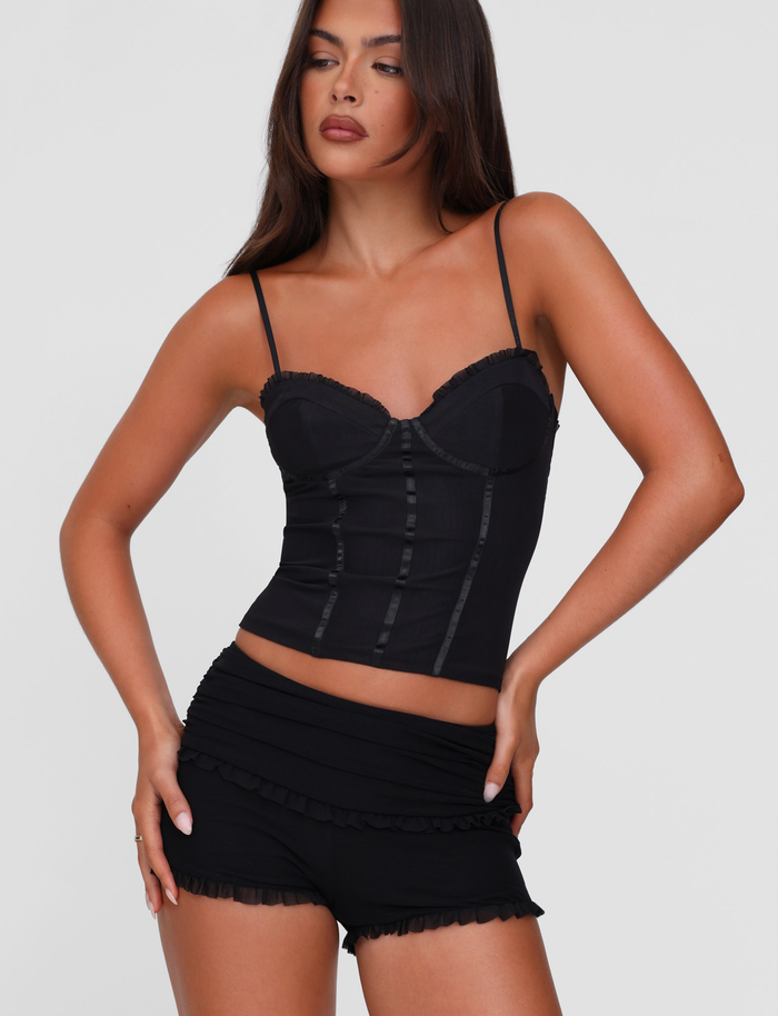 A woman with long brown hair poses in TIGERMIST's GINNY SHORT - BLACK mini shorts with ruffle details and a fitted black camisole, standing against a plain light background.