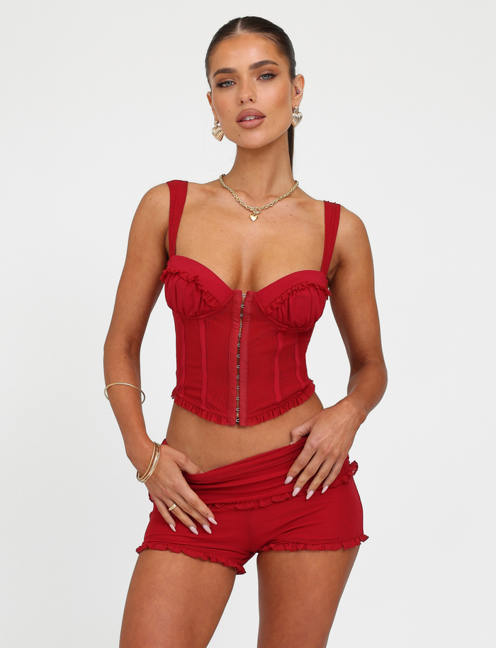 A woman poses against a white background wearing the TIGERMIST GINNY SHORT - RED : CHERRY RED set, featuring a matching red corset top and shorts with a bodycon fit. She accessorizes with gold jewelry and her hair is tied back, hand on waist.