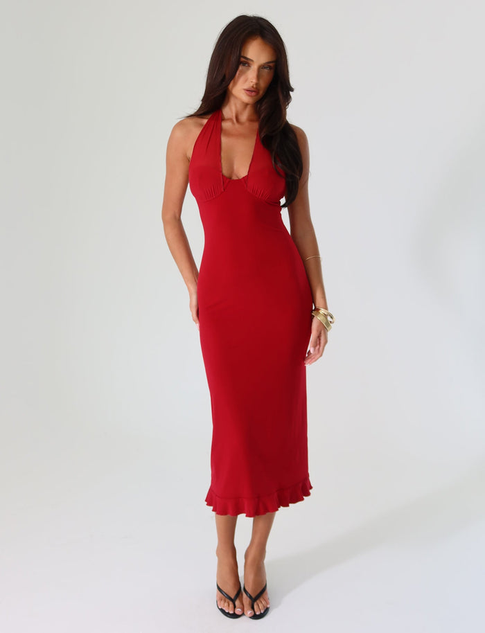 A person with long dark hair is wearing the TIGERMIST GRANGER MIDI DRESS in cherry red, featuring a sleeveless, halter-neck design with a ruffled hem. They are standing against a plain white background and are accessorized with black high-heeled sandals and a gold bracelet.