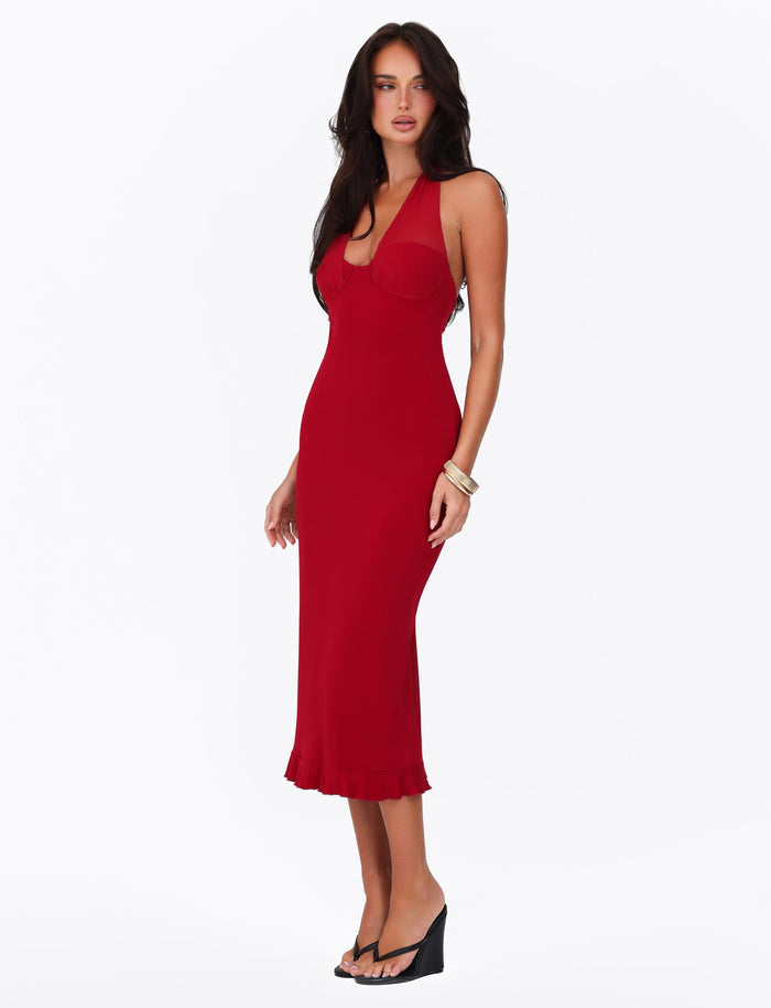 A woman wearing the TIGERMIST GRANGER MIDI DRESS in Cherry Red confidently poses against a white background. She accessorizes her look with black wedge heels and a light-colored bracelet, her dark hair cascading down her shoulders.