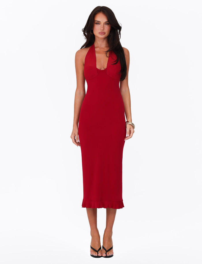 A woman is wearing the TIGERMIST GRANGER MIDI DRESS in CHERRY RED, which is sleeveless, form-fitting, and features a halter neckline. She stands against a plain white background, pairing it with black high-heeled sandals and a bracelet on her left wrist. Her long hair is styled down.