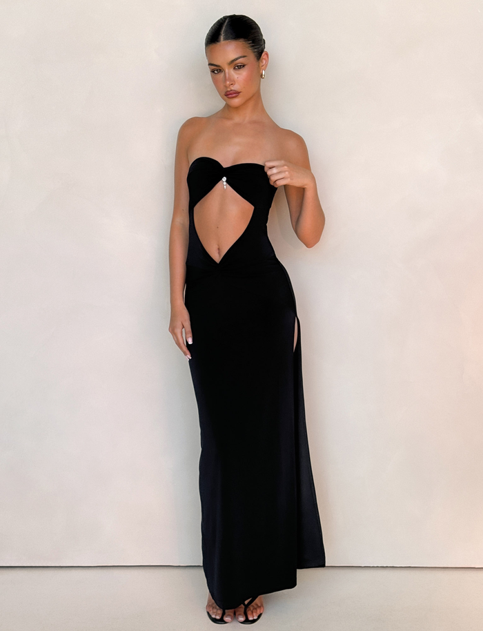 A woman with her hair pulled back and a neutral expression wears the TIGERMIST GRAZIA DRESS - BLACK, a black bandeau maxi dress featuring a midriff cutout and side slit, styled with silver jewel earrings against a plain light wall.