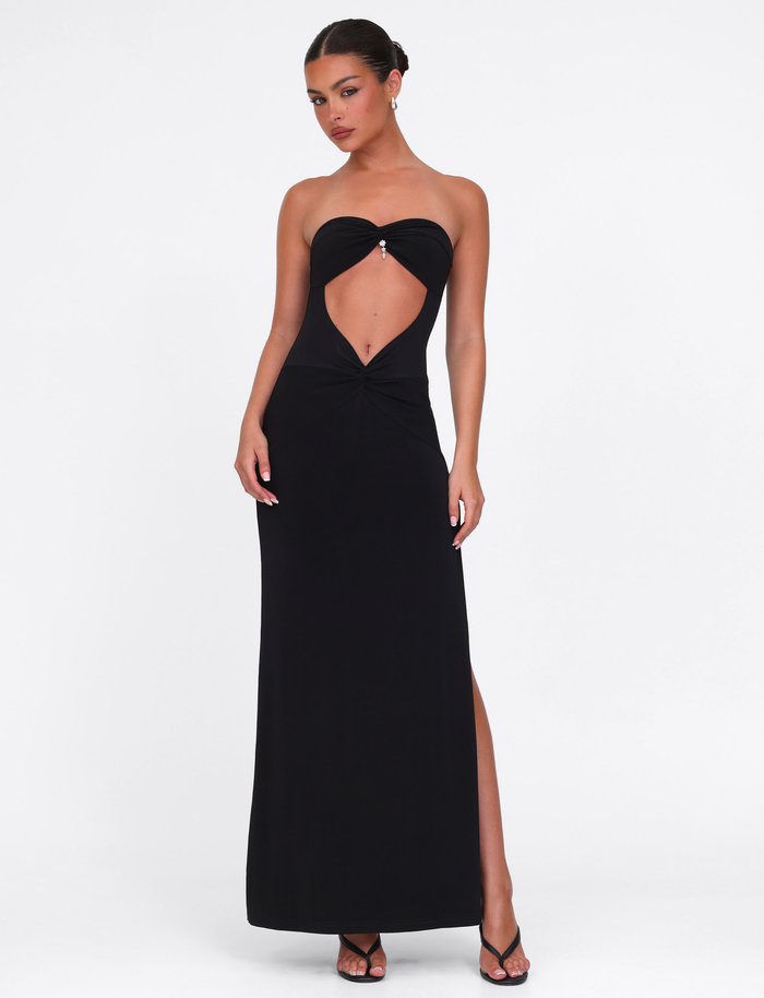 A woman models the TIGERMIST GRAZIA DRESS - BLACK, featuring high stretch fabric, a bandeau neckline, front cutout, and high slit. She pairs it with black high-heeled sandals against a plain white background.