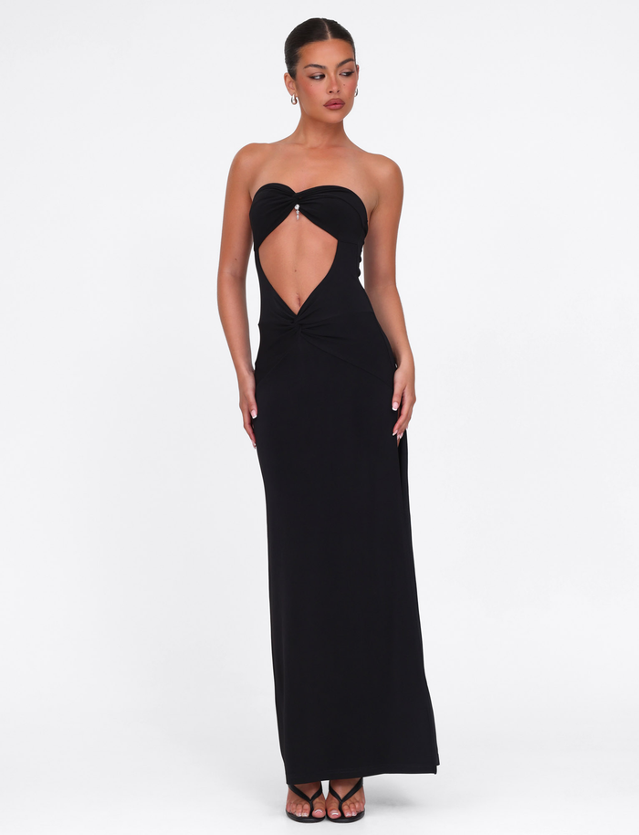 A woman wears the TIGERMIST GRAZIA DRESS - BLACK, a high-stretch bandeau maxi with front cutout and knot detail, styled with slicked-back hair and strappy black heels against a plain white background.