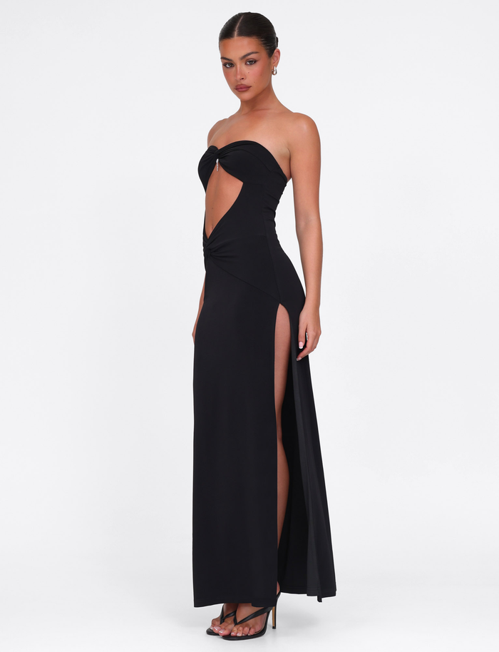 A woman wears the TIGERMIST GRAZIA DRESS - BLACK, featuring a bandeau neckline, front cutout, and high side slit, paired with black heeled sandals. She poses with her hair tied back against a plain white background.