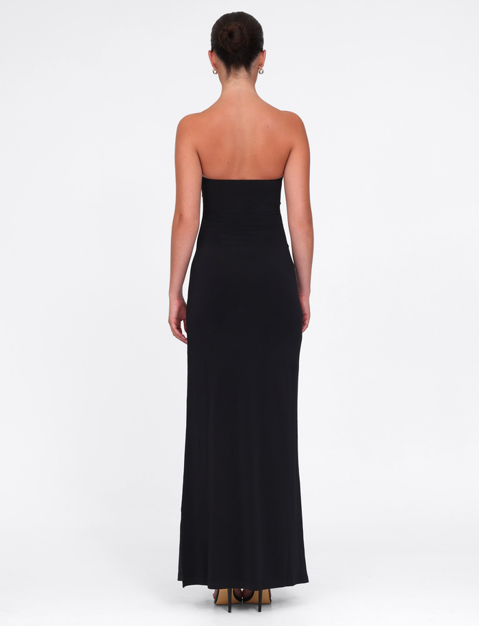 A woman faces away from the camera in the TIGERMIST GRAZIA DRESS - BLACK, a high stretch fabric bandeau maxi dress, paired with high heels against a plain white background.