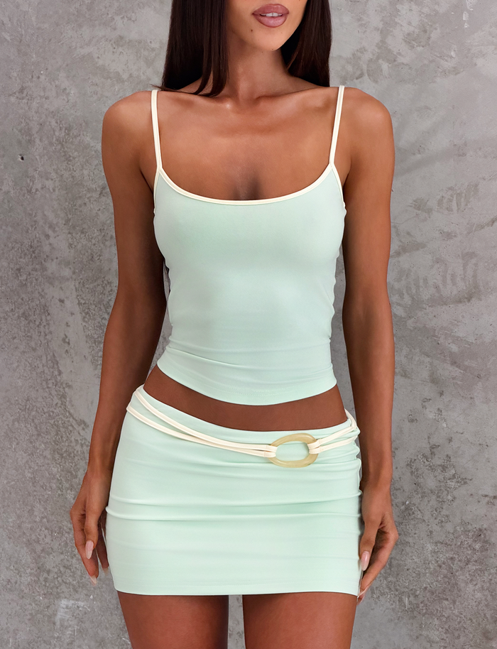 A woman stands against a textured gray background wearing the TIGERMIST GREER MINI SKIRT in mint green and a pastel green fitted tank top with thin straps, paired with a cream belt featuring a circular buckle.