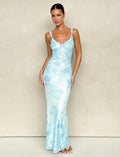 A person wearing the GUILIANNA FRILL MAXI DRESS - BLUE : FLORAL : TAPESTRY from TIGERMIST poses against a plain beige background. They have long hair tied back and are standing with one hand on their hip, highlighting the frill shoulder strap, while wearing white sandals.