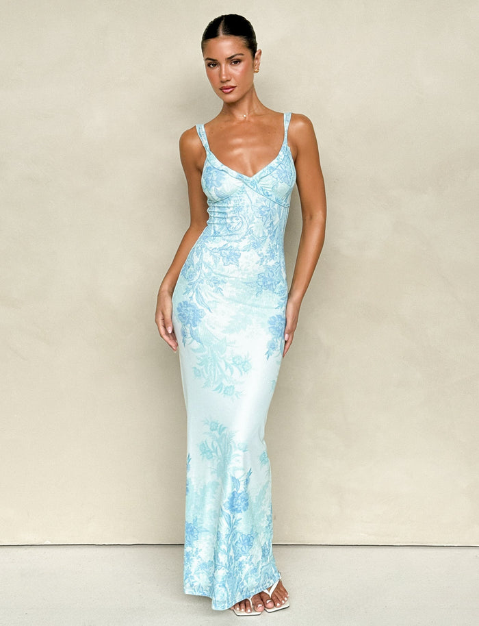 A person wearing the GUILIANNA FRILL MAXI DRESS - BLUE : FLORAL : TAPESTRY from TIGERMIST poses against a plain beige background. They have long hair tied back and are standing with one hand on their hip, highlighting the frill shoulder strap, while wearing white sandals.