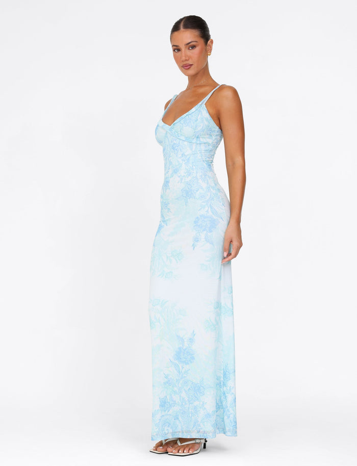 A woman wearing the TIGERMIST GUILIANNA FRILL MAXI DRESS in a light blue floral tapestry pattern stands against a plain white background. The dress features frill shoulder straps, which she complements with strappy white heels and her hair styled in a tied-back manner. Her pose is relaxed, facing slightly to the side.