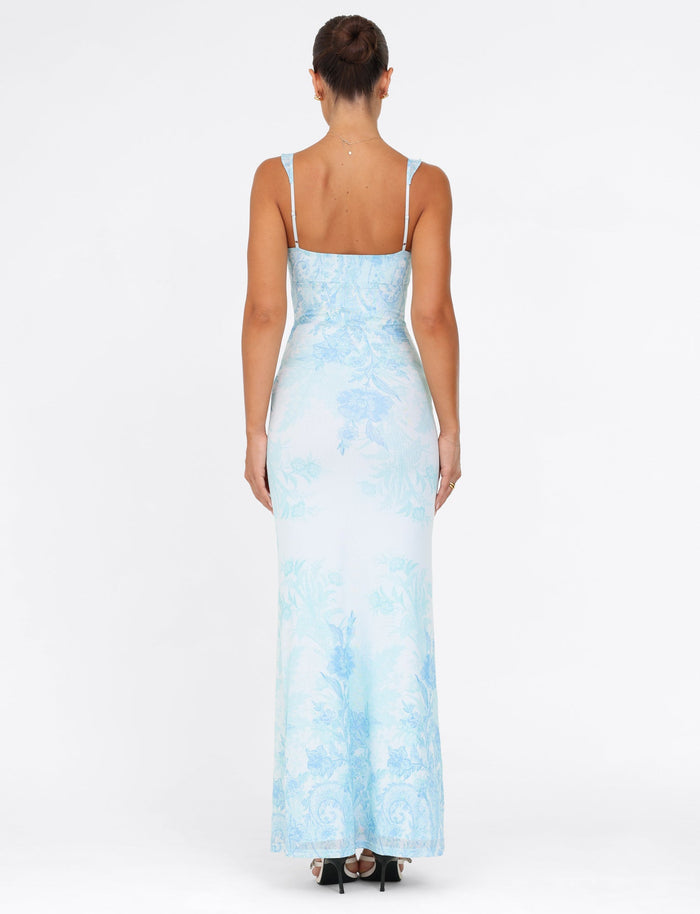 A woman is seen wearing the GUILIANNA FRILL MAXI DRESS from TIGERMIST, featuring a fitted silhouette with a blue floral tapestry pattern and frill shoulder straps. The dress showcases thin straps and a straight neckline. Her hair is styled in an updo, and she pairs the look with black heels against a plain white background.