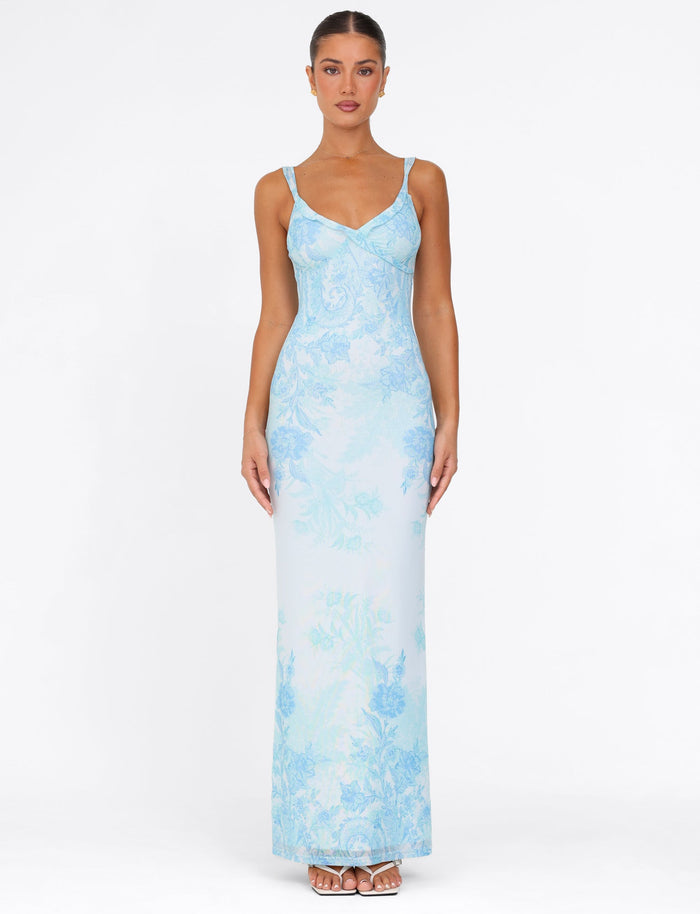 A woman stands against a plain white background, showcasing the TIGERMIST Guilianna Frill Maxi Dress in blue floral tapestry. Her hair is pulled back, and she complements the dress with white sandals.