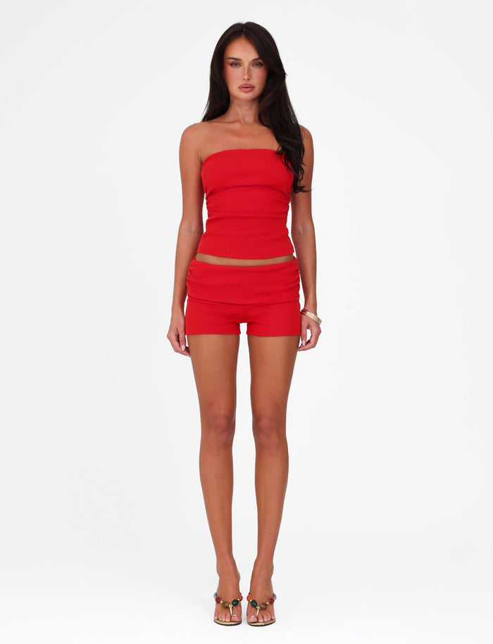 Against a white background, a woman wears the HARRIETT RIB TUBE TOP - RED from TIGERMIST, along with matching shorts. Her long dark hair cascades over her shoulders, and she completes the chic bodycon look with high-heeled sandals.