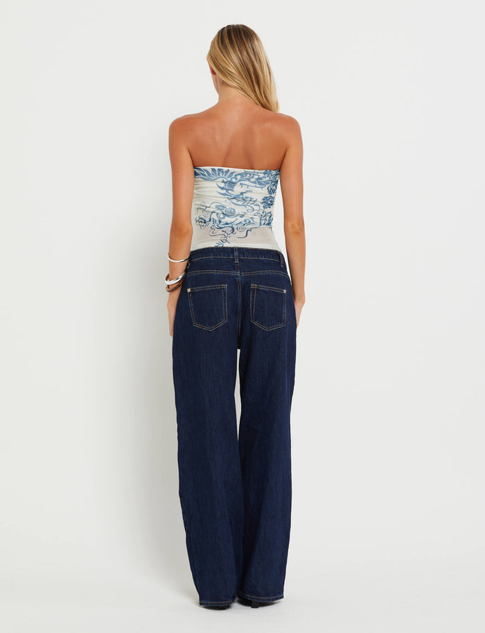 A blonde woman stands with her back to the camera, showcasing the TIGERMIST HARRIETT TUBE TOP - BLUE : TATTOO : TATTOO PRINT paired with dark blue wide-leg jeans. Her arms are adorned with several bracelets against a plain white background.