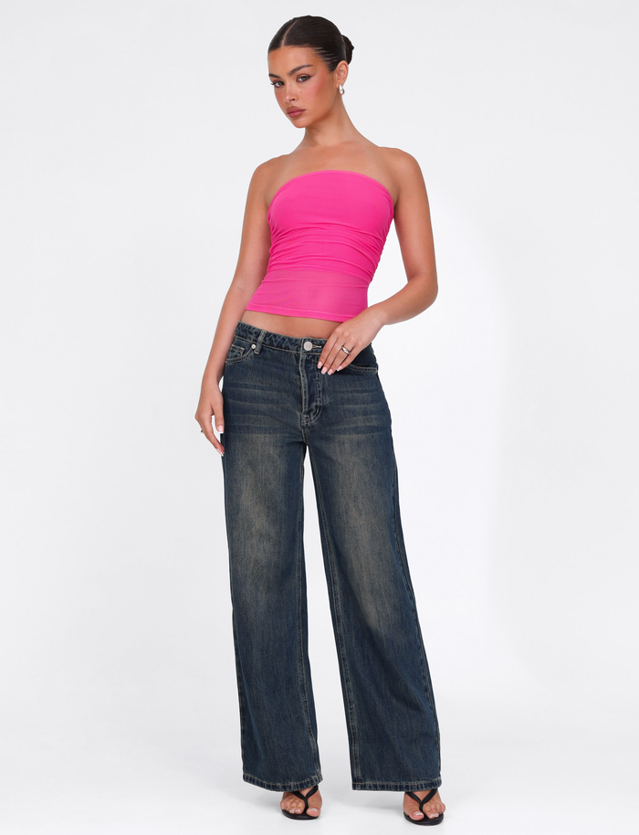 A woman stands against a white background, wearing the TIGERMIST HARRIETT TUBE TOP in pink, bodycon style, paired with wide-leg blue jeans and black heeled sandals. Her hair is tied back and she poses with one hand on her hip.