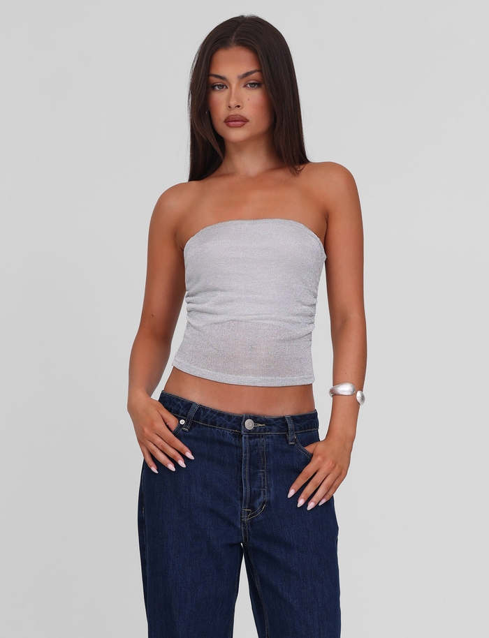 A woman with long brown hair wears the TIGERMIST HARRIETT TUBE TOP in silver sparkle jersey and dark blue jeans, standing against a plain light gray background with one hand on her hip.