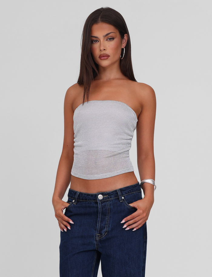 A woman with straight dark hair wears the TIGERMIST HARRIETT TUBE TOP in silver, paired with dark blue jeans, silver hoop earrings, and a silver bangle. She stands against a light gray background with her hands in her pockets.