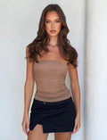 A young woman with long, wavy brown hair stands against a plain white background. Dressed in a chic TIGERMIST HARRIETT TUBE TOP - TAN and a black mini skirt, her expression remains neutral as her arms rest loosely at her sides.
