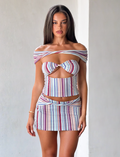 A woman with long straight hair wears the TIGERMIST HAZE TWIST SKIRT - VARIEGATED STRIPE and a matching off-shoulder crop top, featuring multicolored vertical stripes, standing against a light-colored wall.