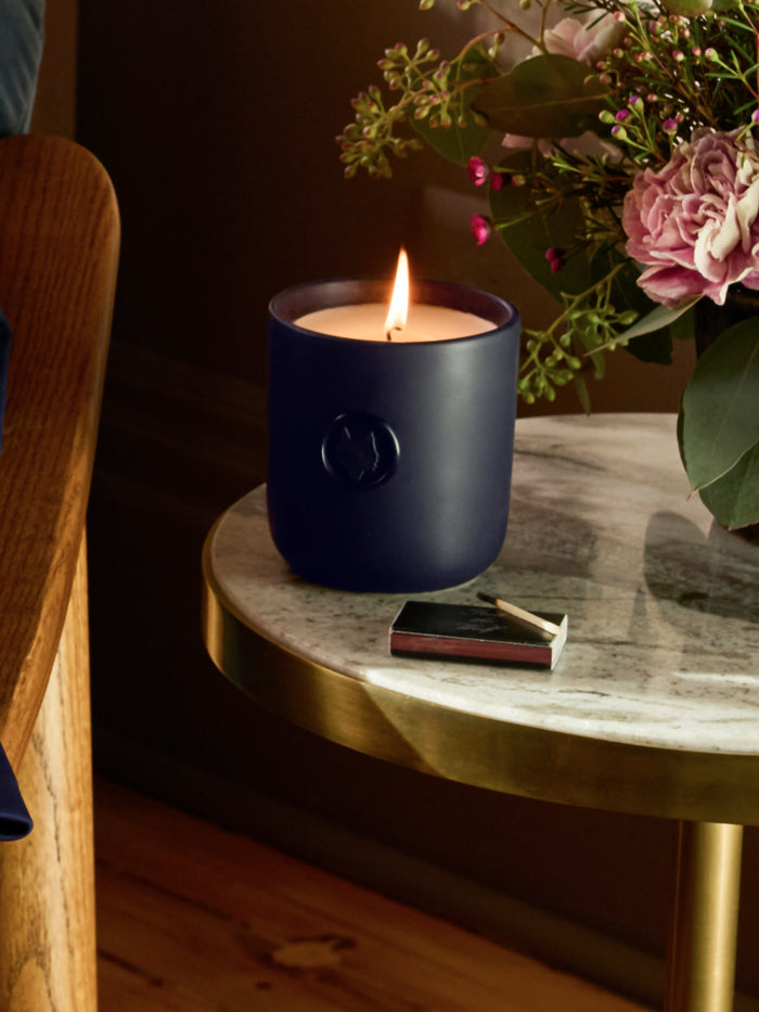 Greyson Icon Wolf Fig Scented Candle burning on marble side table with flowers and matchbook - Lifestyle | Greyson Clothiers