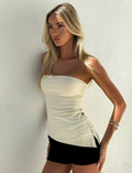 A woman with long blonde hair wears the TIGERMIST INDIRA TOP in cream, a strapless fitted piece, paired with black shorts. She stands against a white wall, gazing sideways with a serious look, her bent arm accessorized by two silver bangles.