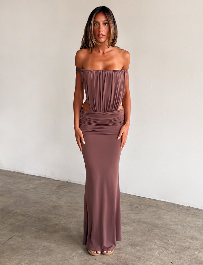 A woman stands indoors by a plain wall, wearing the TIGERMIST IOLLA STRAPLESS MAXI DRESS in mocha, featuring a strapless ruched bodice and waist cutouts. She has straight shoulder-length hair and neutral sandals.
