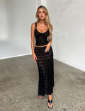 A woman with long blonde hair is standing on a polished concrete floor in a minimalist room. She is wearing a matching two-piece black lace set from Tigermist, featuring the ISADORA SKIRT - BLACK : LACE and a sleeveless crop top. She completes the look with black sandals.