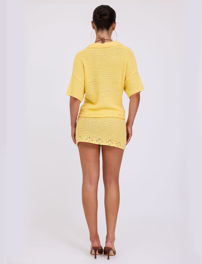 A person with dark hair styled in a bun stands facing away, showcasing the TIGERMIST JANAI TOP - YELLOW : LEMON with short sleeves and a matching yellow mini skirt. They are also wearing high-heeled shoes and large hoop earrings. The background is plain and white.