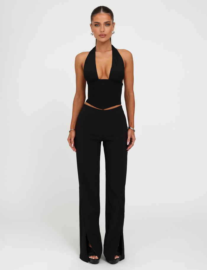 A woman faces forward against a white background, wearing the TIGERMIST JANECE TOP in black with high-waisted black pants featuring front slits, styled with earrings, bracelets, and rings.