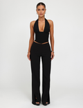 A woman faces forward against a white background, wearing the TIGERMIST JANECE TOP in black with high-waisted black pants featuring front slits, styled with earrings, bracelets, and rings.
