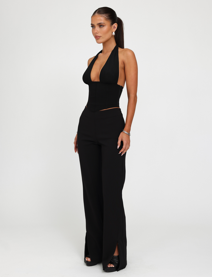 A woman wears the TIGERMIST JANECE TOP - BLACK, a sleeveless halter corset with a deep neckline, paired with high-waisted black pants featuring front hem slits. Her hair is in a ponytail and she completes the look with black open-toe heels.