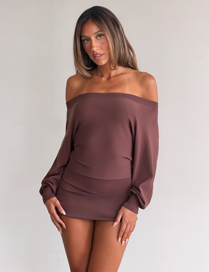A woman with straight, shoulder-length hair wears the TIGERMIST JANINE MINI DRESS in chocolate—an off-shoulder, long-sleeve mini—while standing against a plain light-colored wall and looking at the camera with a relaxed expression.