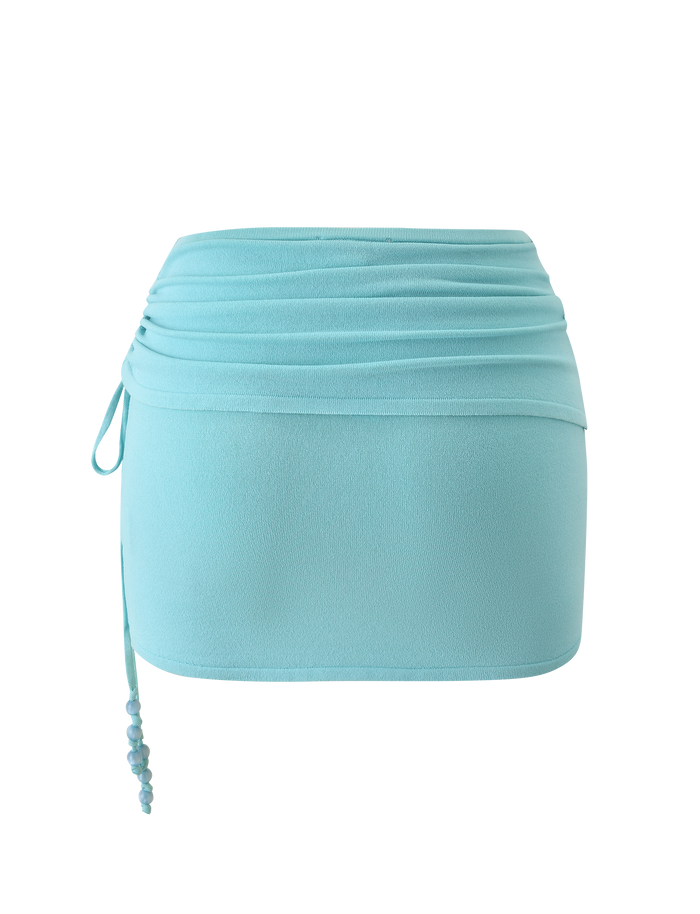 The JASMINA KNIT MINI SKIRT - BLUE by TIGERMIST features a light blue hue, ruched waistband, side drawstring with beaded detail, and a fitted silhouette for a smooth, effortlessly stylish look.