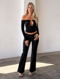 A woman with long blonde hair models the TIGERMIST JENESIS LONG SLEEVE TOP - BLACK, featuring an off-the-shoulder cutout. She pairs it with black pants and heeled sandals, posing confidently against a white wall with one hand on her hip.