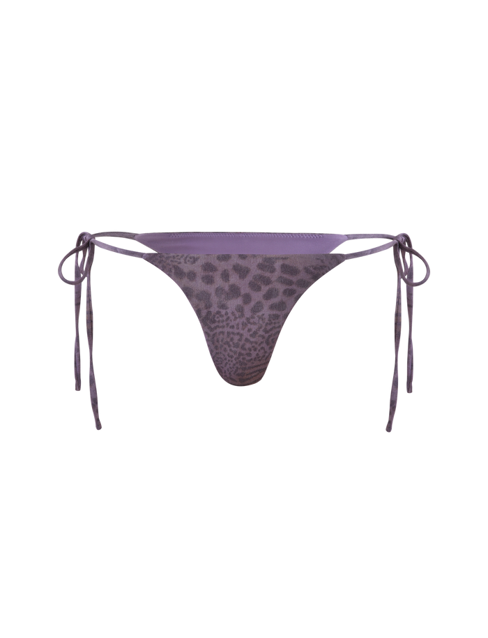 The JESSALYN BIKINI BOTTOM - PURPLE TIGER by TIGERMIST features a purple sparkle animal print, side tie strings, and cheeky minimal coverage, displayed against a light purple background.