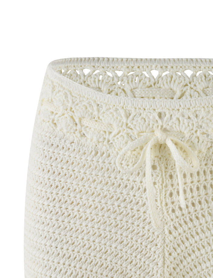 A close-up of the JONAS SHORT - WHITE : CREAM from TIGERMIST showcases intricate crochet patterns with a decorative knit tie at the mid-rise waist. The texture and weave details are prominently visible, highlighting the high stretch fabric's flexibility.