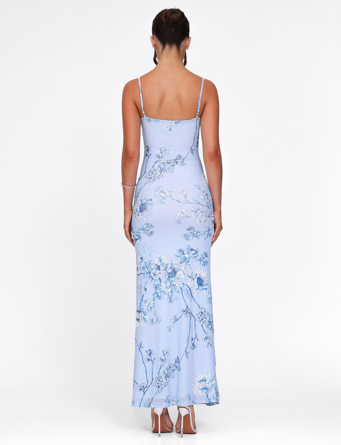 A woman faces away, wearing the TIGERMIST JULIANNA DRESS in Blue Ceramic Floral—a light blue, sleeveless maxi with thin straps. Her hair is tied up and she pairs it with white heeled sandals against a white background.